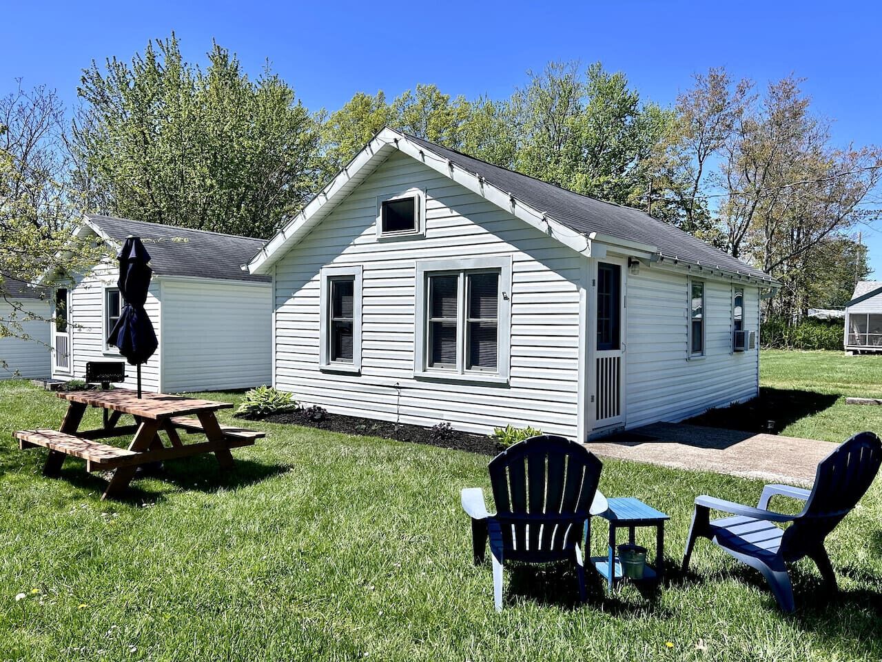 A small white cabin with a pitched roof, a picnic table, and two Adirondack chairs in the yard.