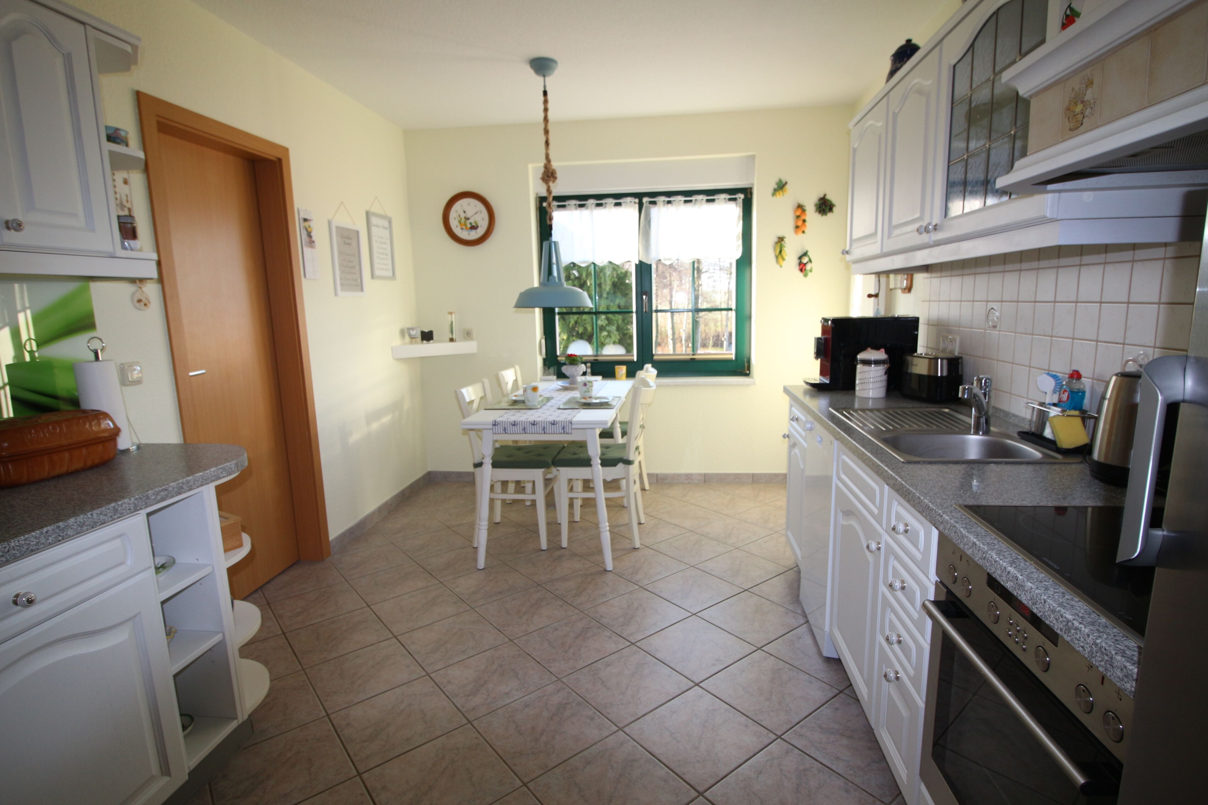 Une cuisine avec des meubles blancs, une table à manger avec des chaises, une fenêtre avec des rideaux et un évier.