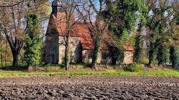 Eine steinerne Kirche mit rotem Dach, umgeben von Bäumen und einem Ackerland.
