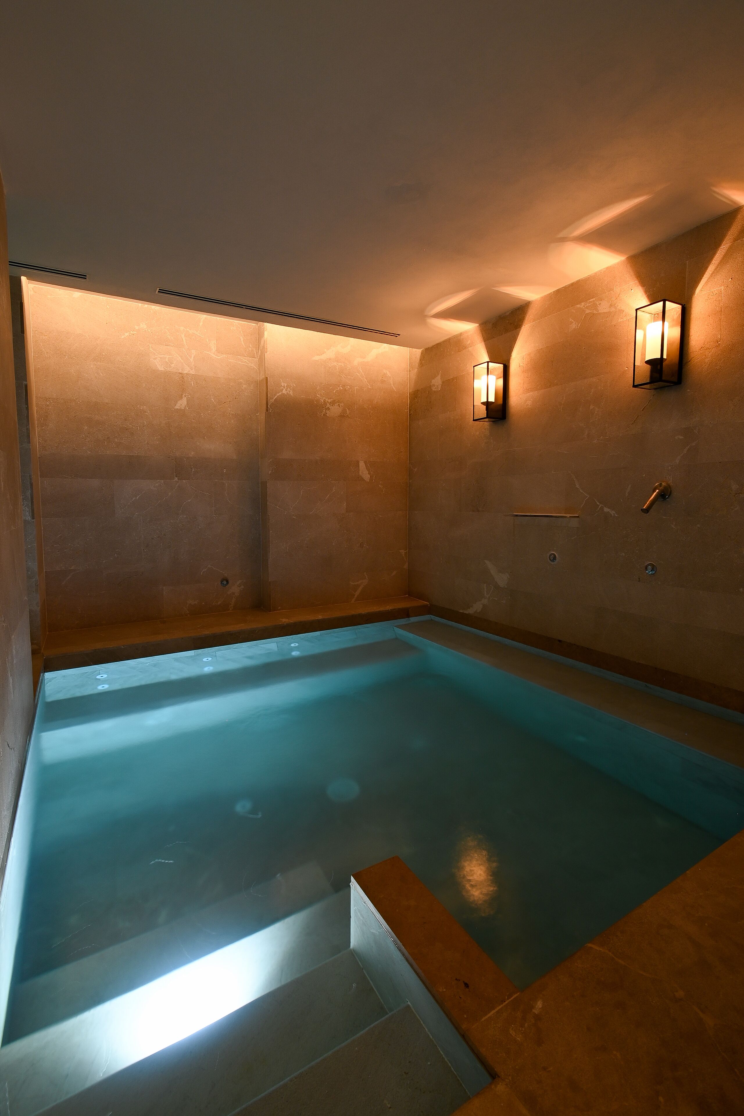Une salle de spa dotée d’une piscine rectangulaire, de luminaires fixés au mur et d’un mur recouvert de carreaux de marbre.