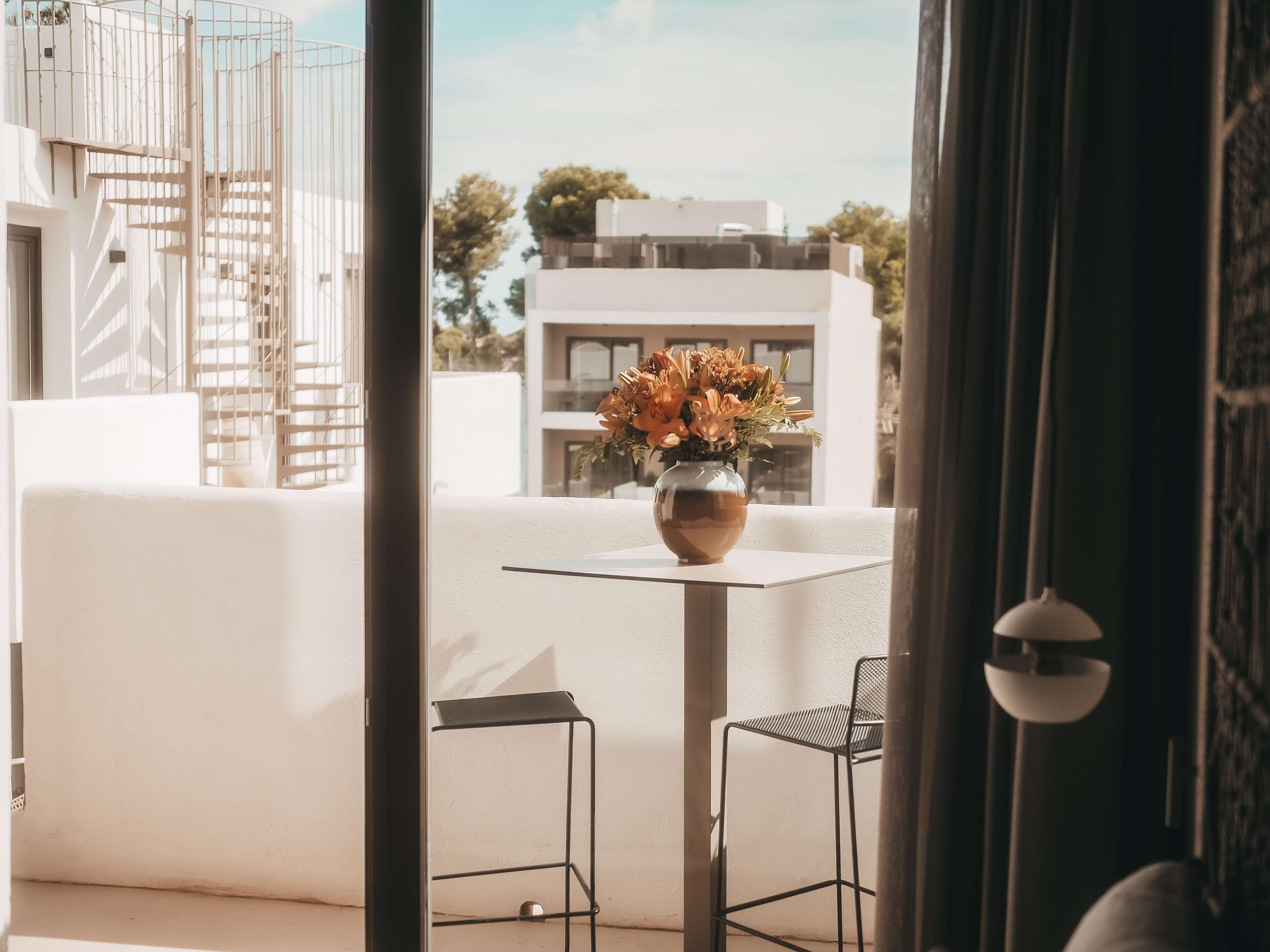 A balcony with a white wall, a metal chair, a metal table, and a vase with flowers.