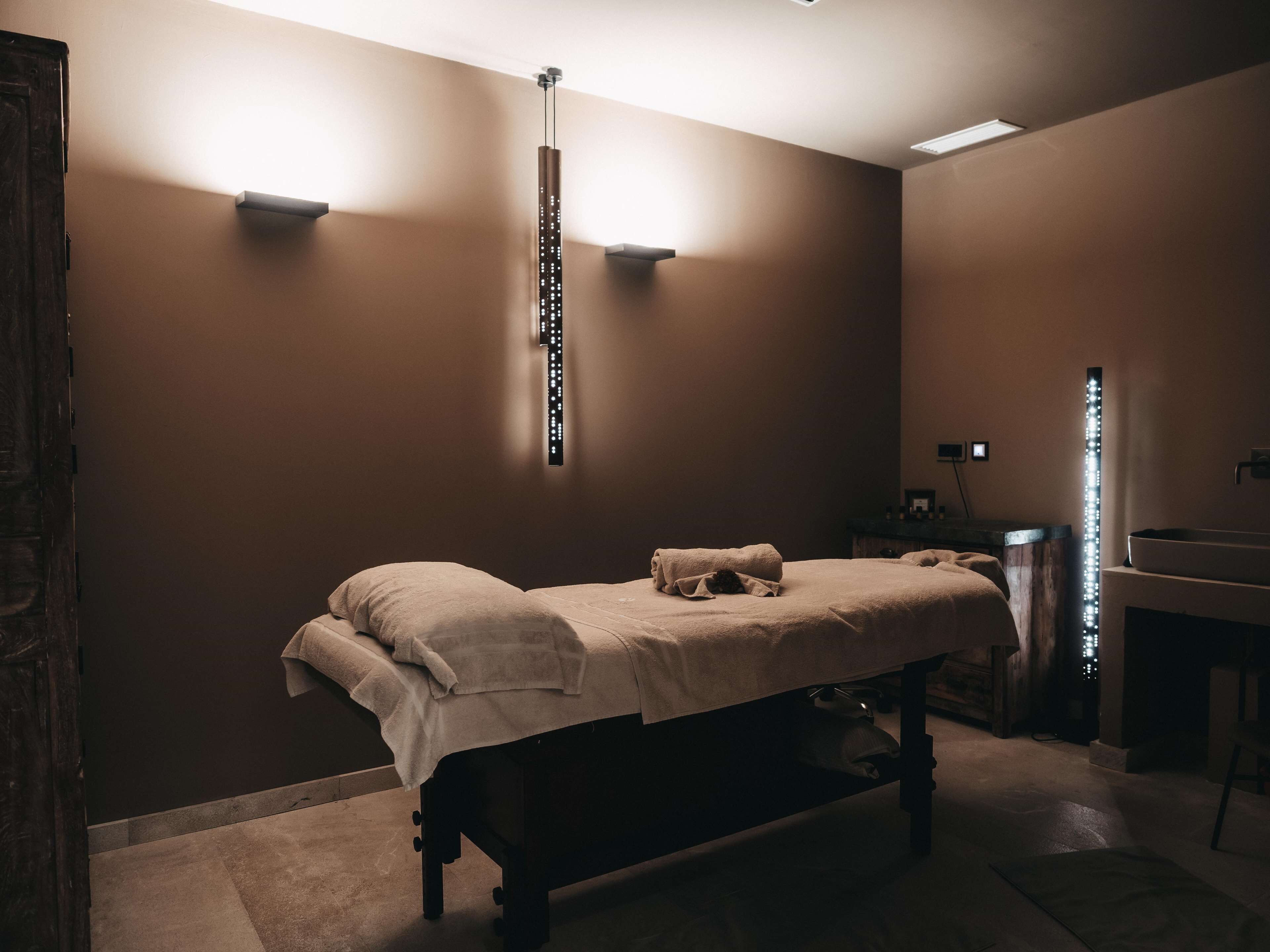 A dimly lit room with a single bed, a small table, and a wall-mounted light fixture.