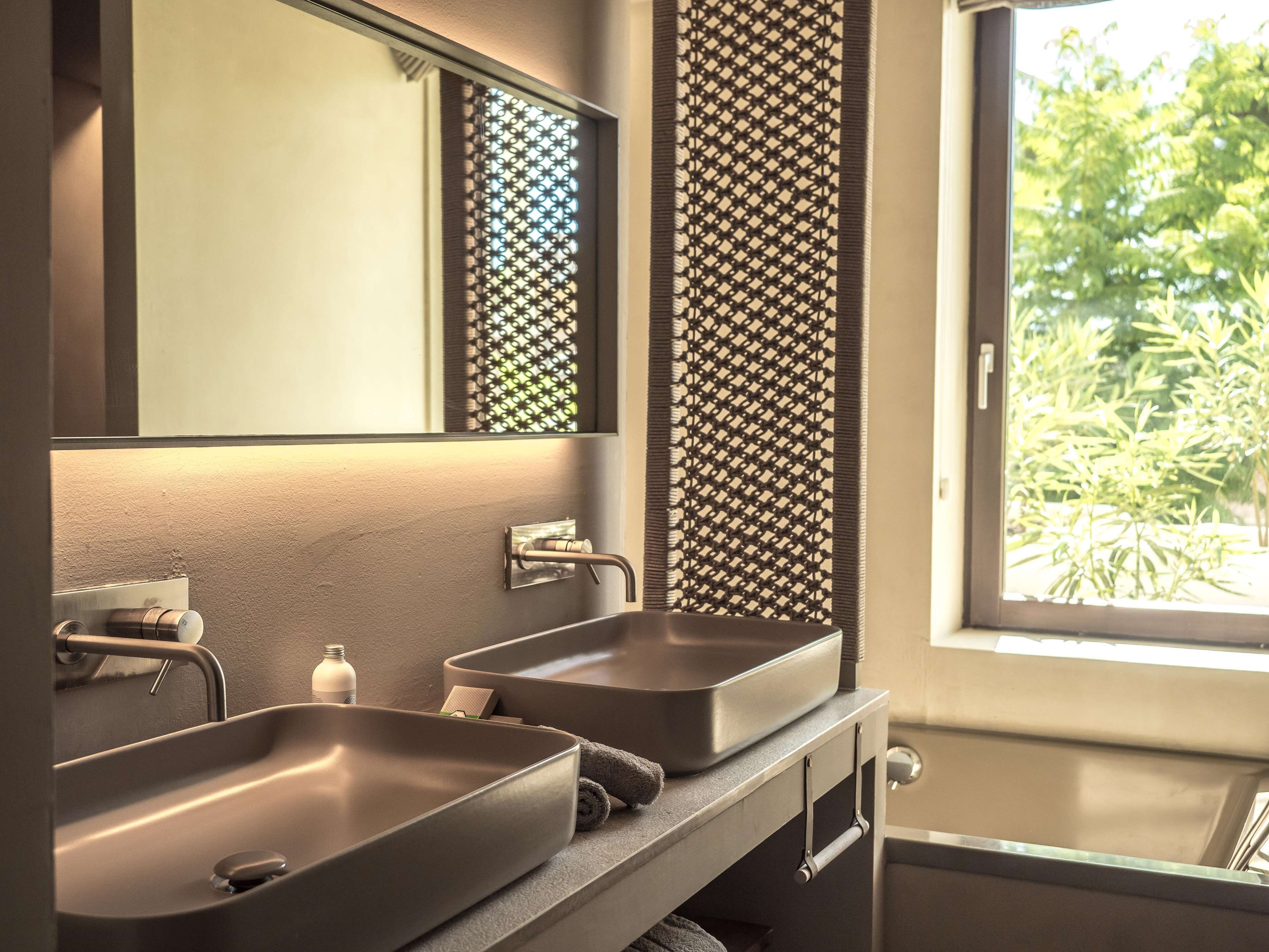 A modern bathroom with two sinks, a large mirror, and a window with a view of greenery.