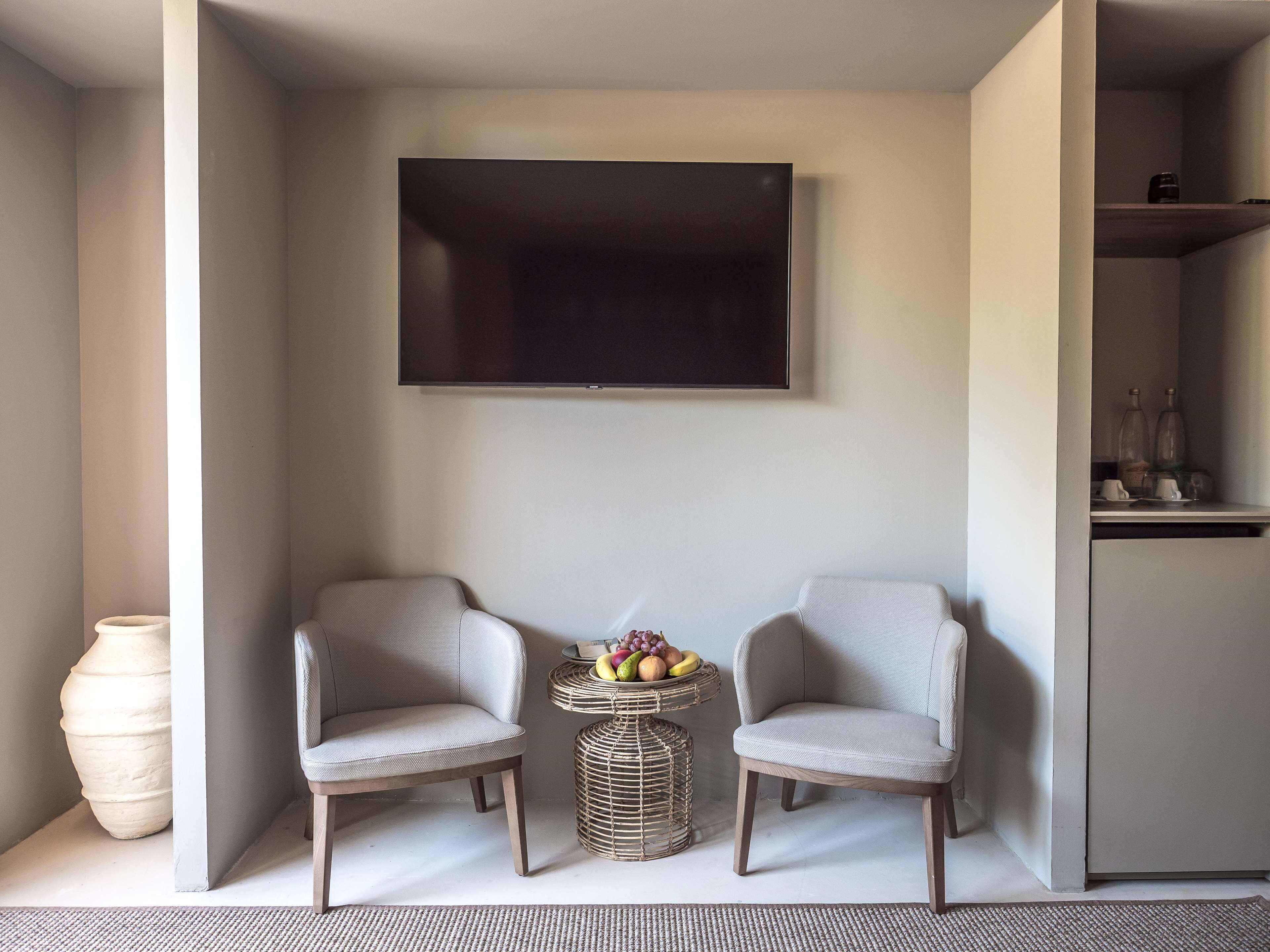A modern living room with two armchairs, a flat-screen TV, a wicker side table with fruit, and a large vase.
