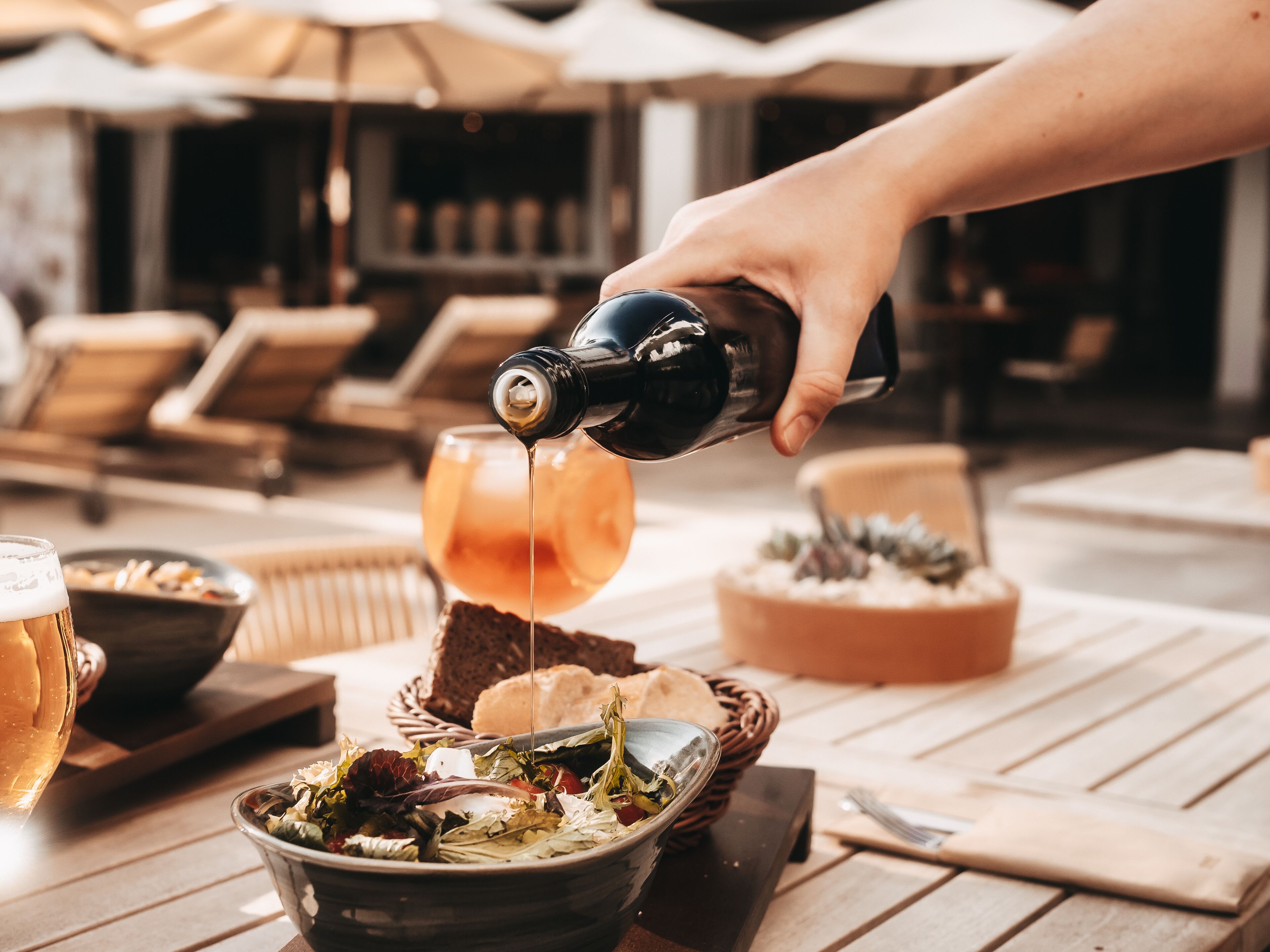 Une main verse une boisson dans un verre posé sur une table en bois, à côté d’un saladier, d’une miche de pain et d’un panier de pain.