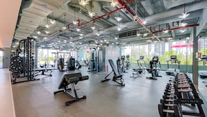 Een fitnessruimte met diverse trainingsapparaten, grote ramen en een plafond met blootgelegde leidingen en verlichtingsarmaturen.
