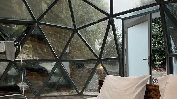 Ein modernes Schlafzimmer mit einer geometrischen Glaswand, einem ordentlich bezogenen Bett und Blick ins Freie.