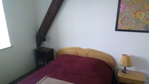 A bedroom with a wooden bed, a red bedspread, a beige blanket, a nightstand with a lamp, and a painting on the wall. - Ecological country house (Èvres)