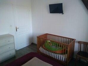 A room with a white door, a small wooden crib with a green mattress, a wooden chair, and a flat-screen TV on the wall. - Ecological country house (Èvres)
