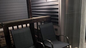 A wooden deck with two chairs and a table, a window with a curtain, and a wall-mounted light fixture. - Rehoboth Beach Condo 1 mile to the beach and Boardwalk w/ pool! Sleeps 6 (Rehoboth Beach)
