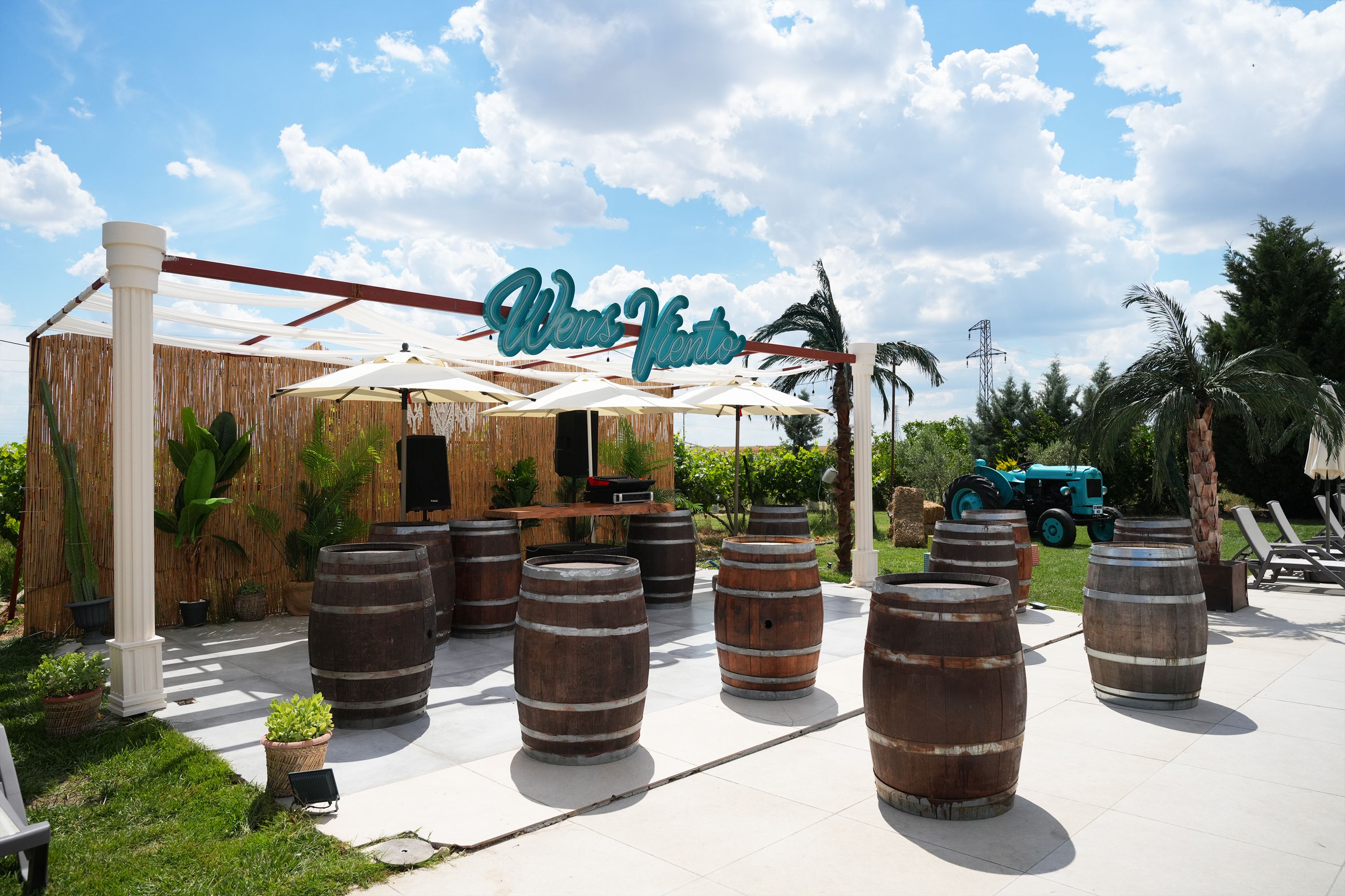 An outdoor seating area with wooden barrels, a sign reading 'Wens Vento,' and a palm tree in the background.