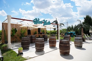 An outdoor seating area with wooden barrels, a sign reading 'Wens Vento,' and a palm tree in the background.