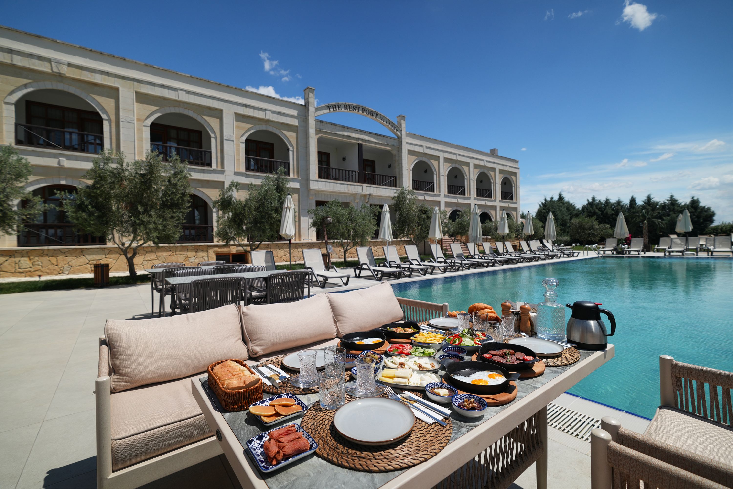 A poolside dining area with a table set for breakfast, overlooking a pool and a building with arched windows.