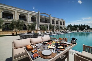 Un área de comedor junto a la piscina con una mesa dispuesta para el desayuno, con vistas a una piscina y un edificio con ventanas arqueadas.