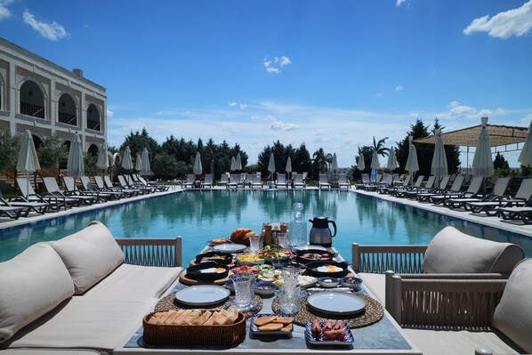 Un área de comedor junto a la piscina con una mesa dispuesta para comer, sillones de descanso y una vista despejada de la piscina.