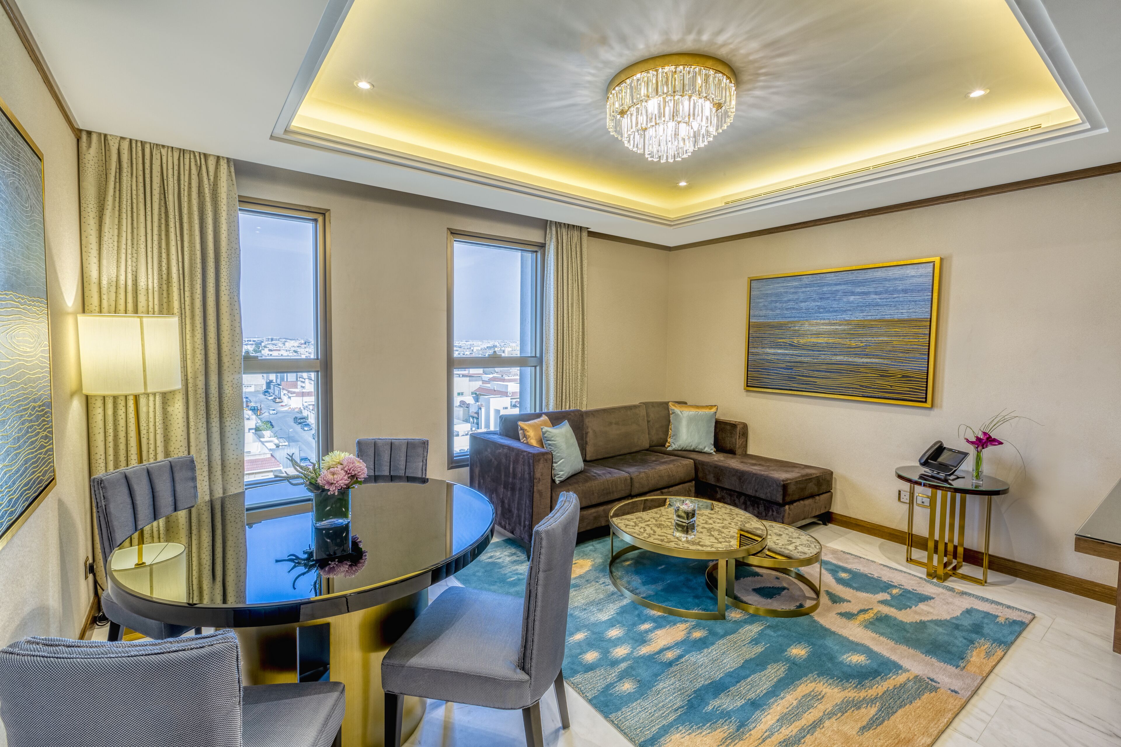 A modern hotel room with a dining area, a sofa set, a chandelier, and a city view.