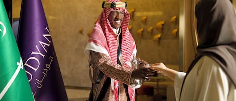 Un homme vêtu d’un costume traditionnel, tenant une tasse, se tient dans une pièce décorée de drapeaux et dotée d’un intérieur moderne.