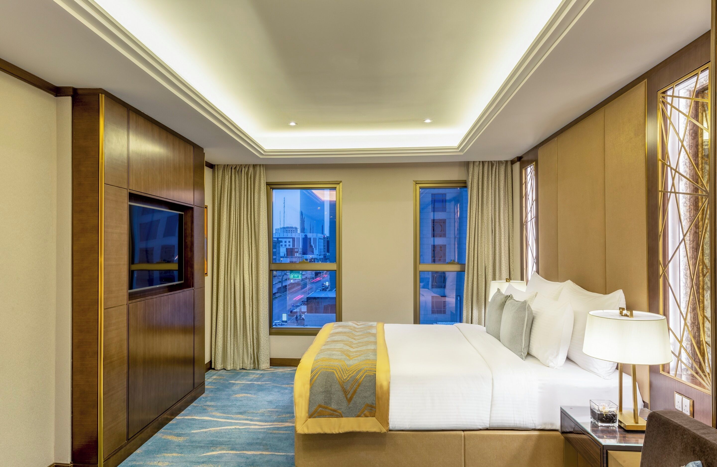 Une chambre d’hôtel avec un lit, une télévision, un bureau avec une lampe et deux fenêtres avec des rideaux.