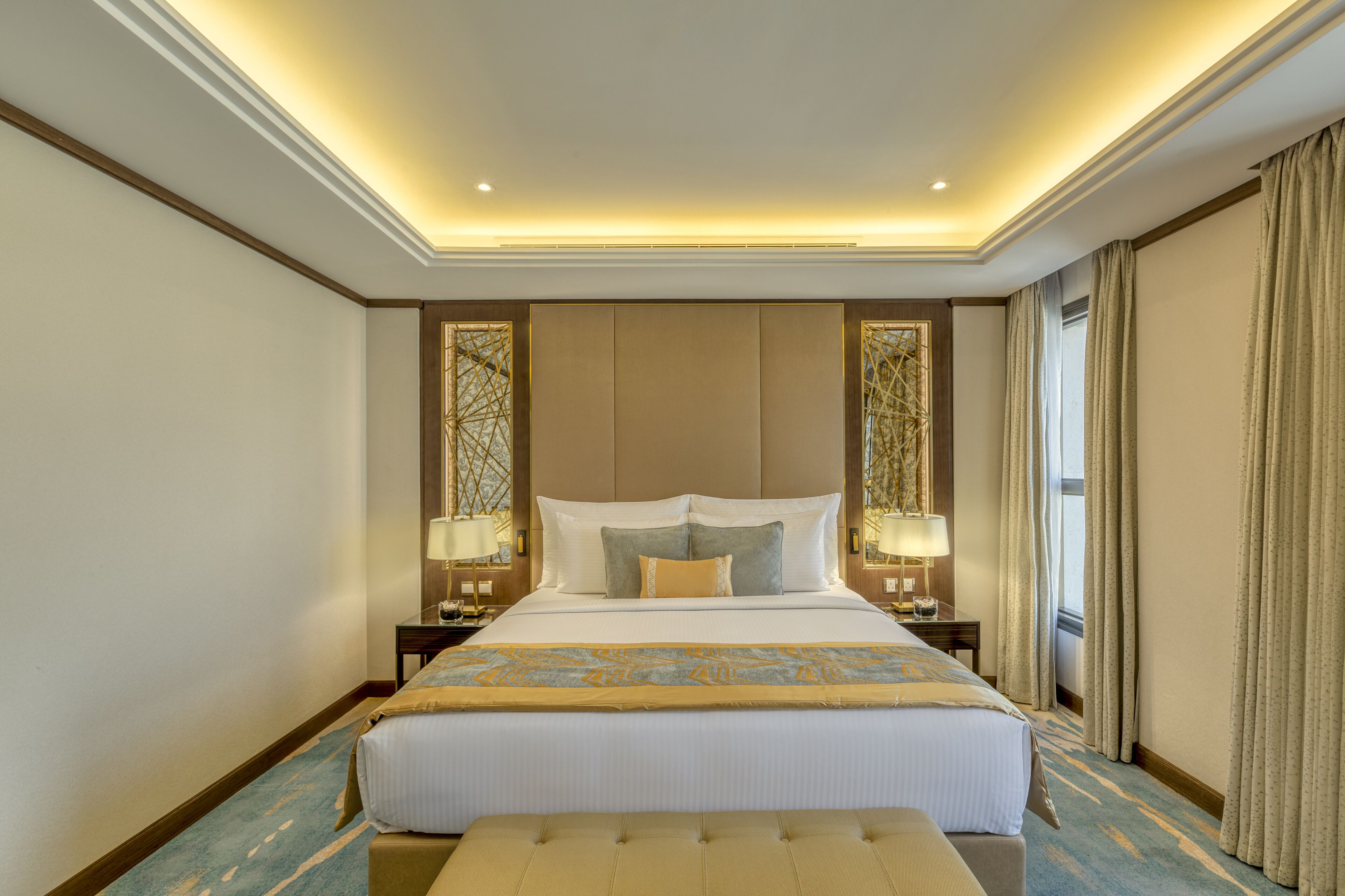 A hotel room with a large bed, two bedside tables with lamps, a window with curtains, and a ceiling with recessed lighting.