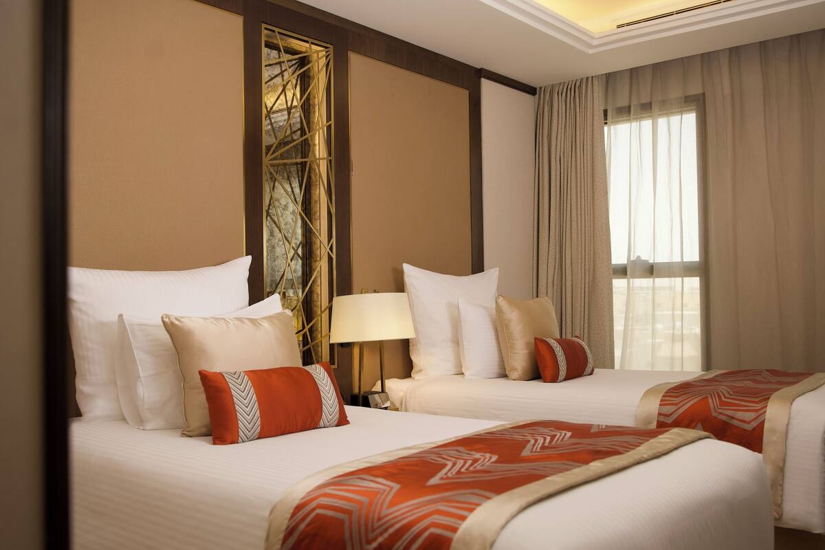 A hotel room with two beds, beige walls, a decorative wall panel, a large window with curtains, and a nightstand with a lamp.