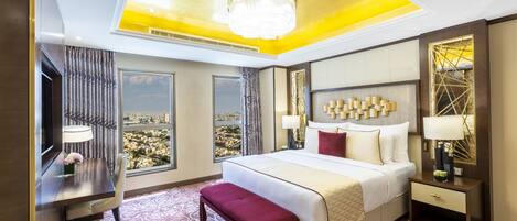 A hotel room with a large bed, a desk, a bench, and a view of a cityscape through large windows.