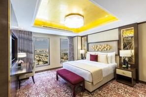 A hotel room with a large bed, a desk, a bench, and a view of a cityscape through large windows.