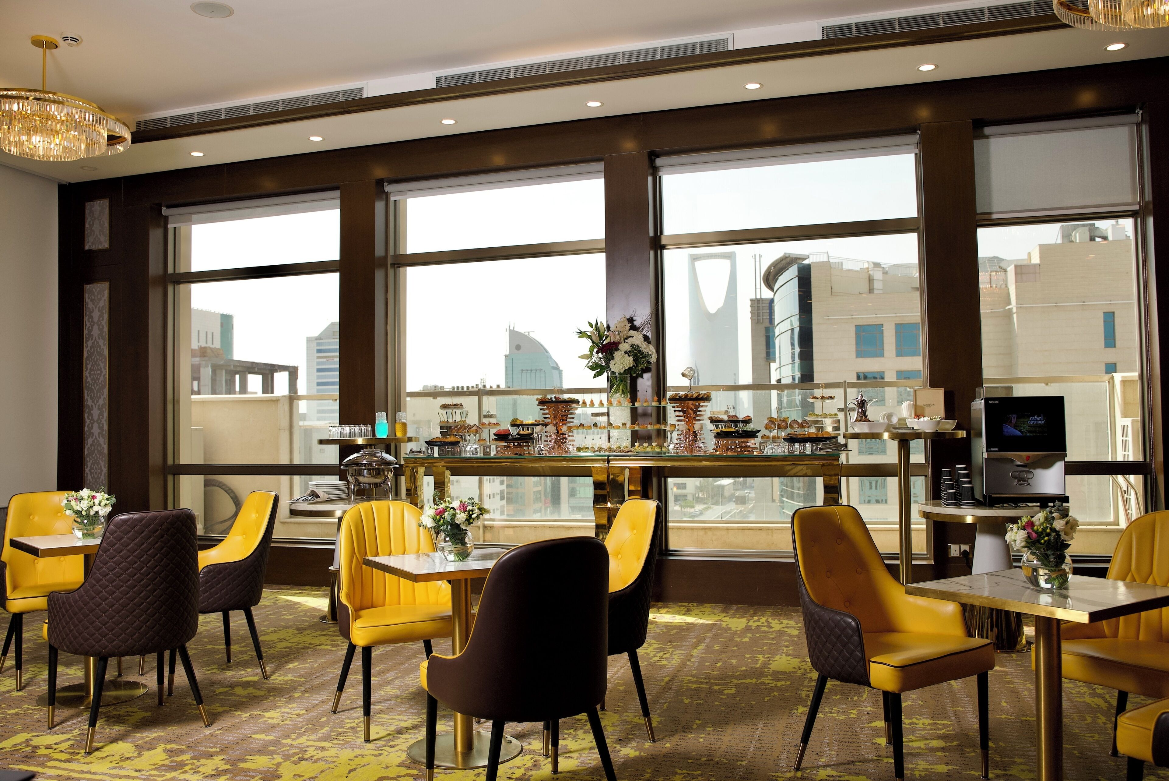 A modern dining area with yellow and black chairs, a bar with a television, and large windows offering a city view.