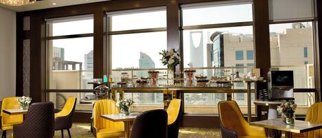 Un espace repas moderne avec des chaises jaunes et noires, un bar équipé d’un téléviseur et de grandes fenêtres offrant une vue sur la ville.