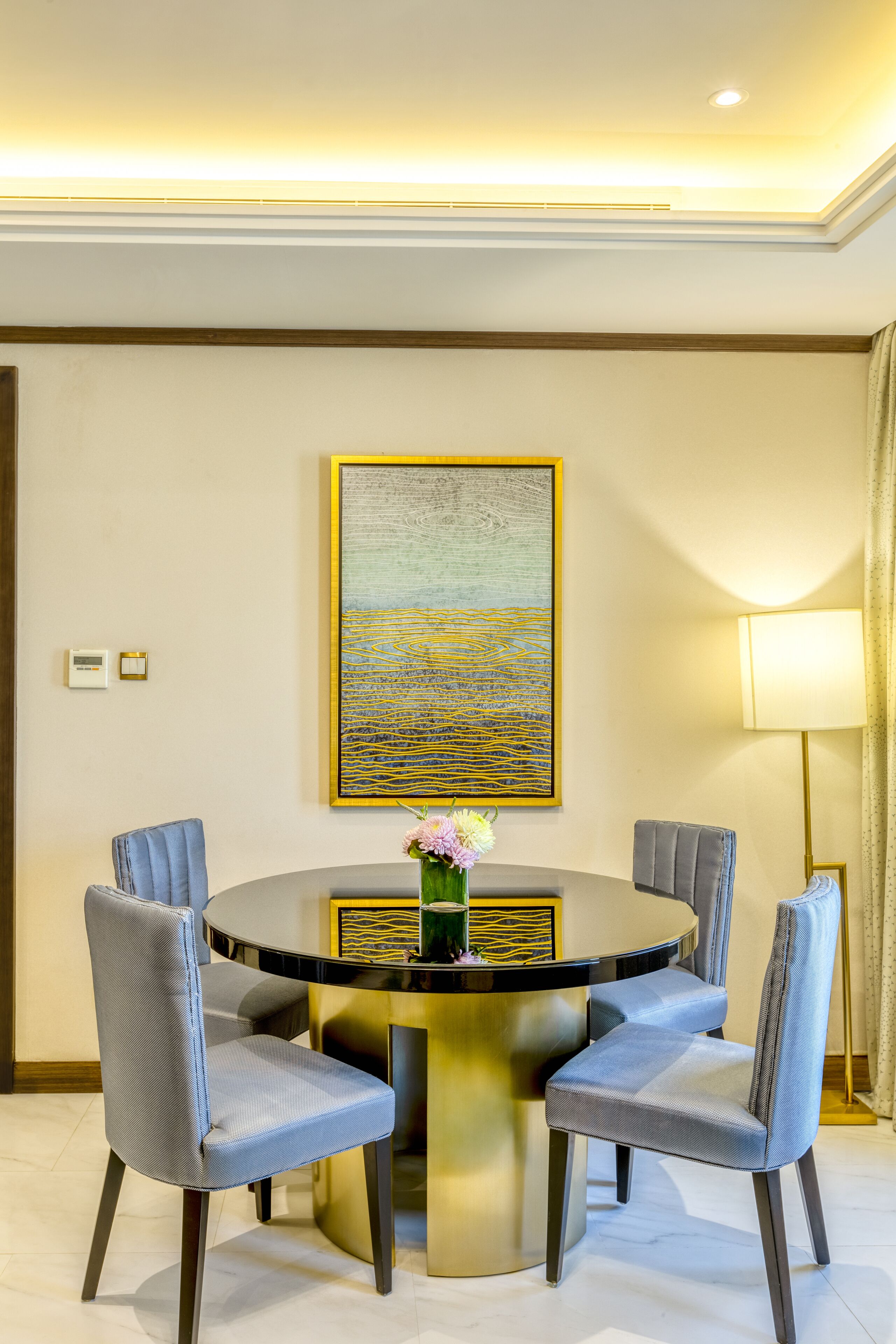 Une table de salle à manger ronde, entourée de quatre chaises rembourrées, un vase rempli de fleurs et, au mur, un tableau abstrait encadré.