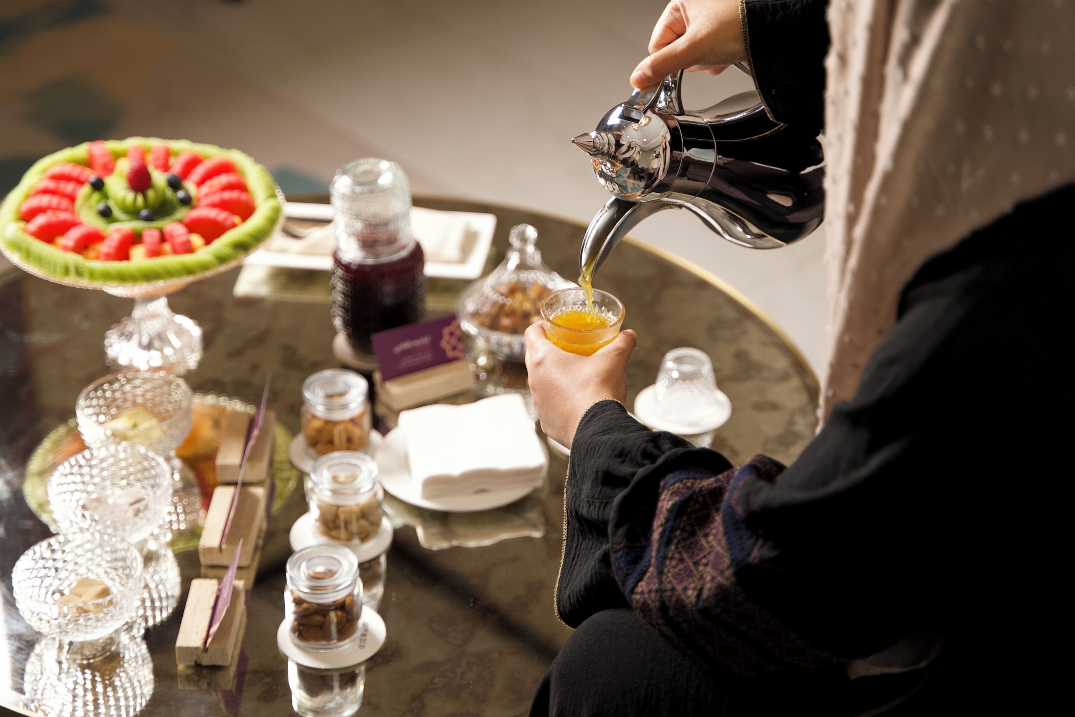 A person pouring a drink into a glass on a table with various food items and condiments.