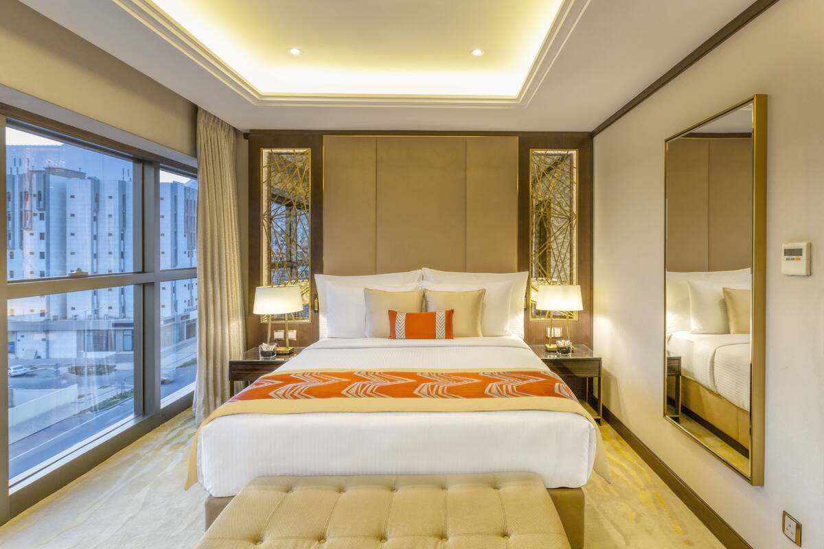 A hotel room with a large bed, two bedside tables with lamps, a large window with a view of buildings, and a mirror on the wall.