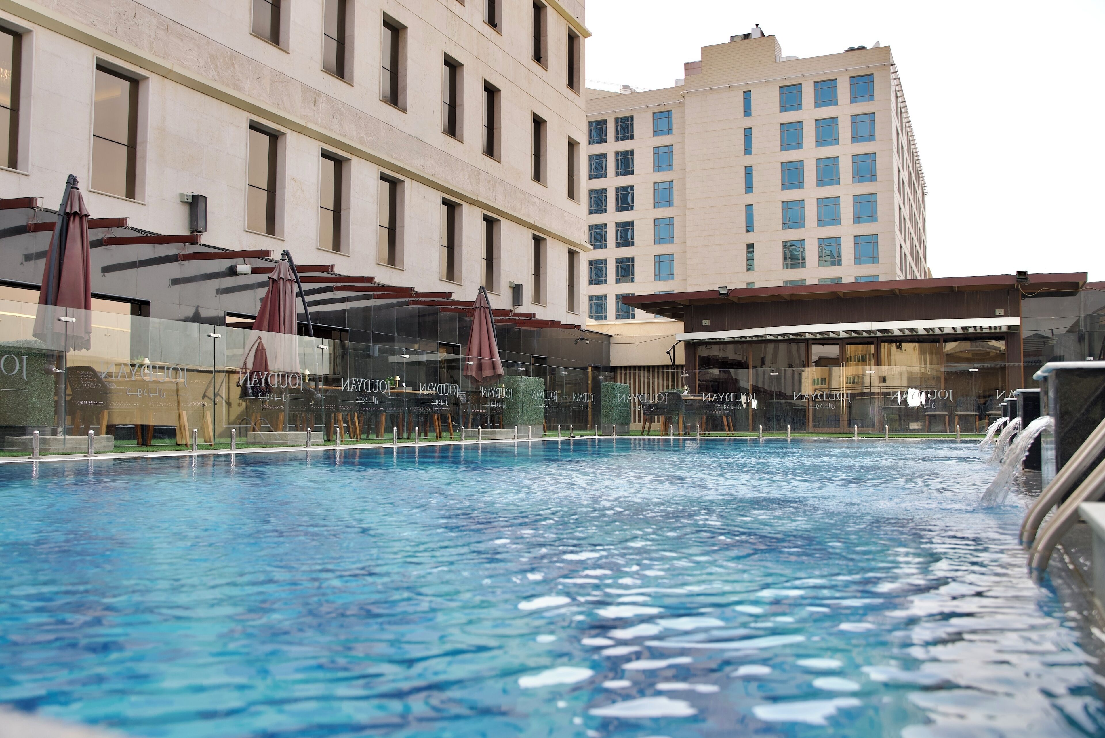 A swimming pool with a glass barrier, surrounded by a building with multiple windows and a separate structure with a roof.