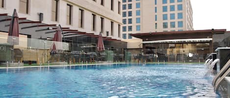 A swimming pool with a glass barrier, surrounded by a building with multiple windows and a separate structure with a roof.