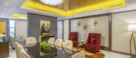 A modern hotel room with a dining area featuring a marble table and upholstered chairs, a red velvet armchair, and a chandelier.