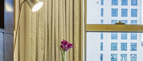 A hotel room with a desk, a chair, a lamp, a vase with a flower, and a view of a building through a window.