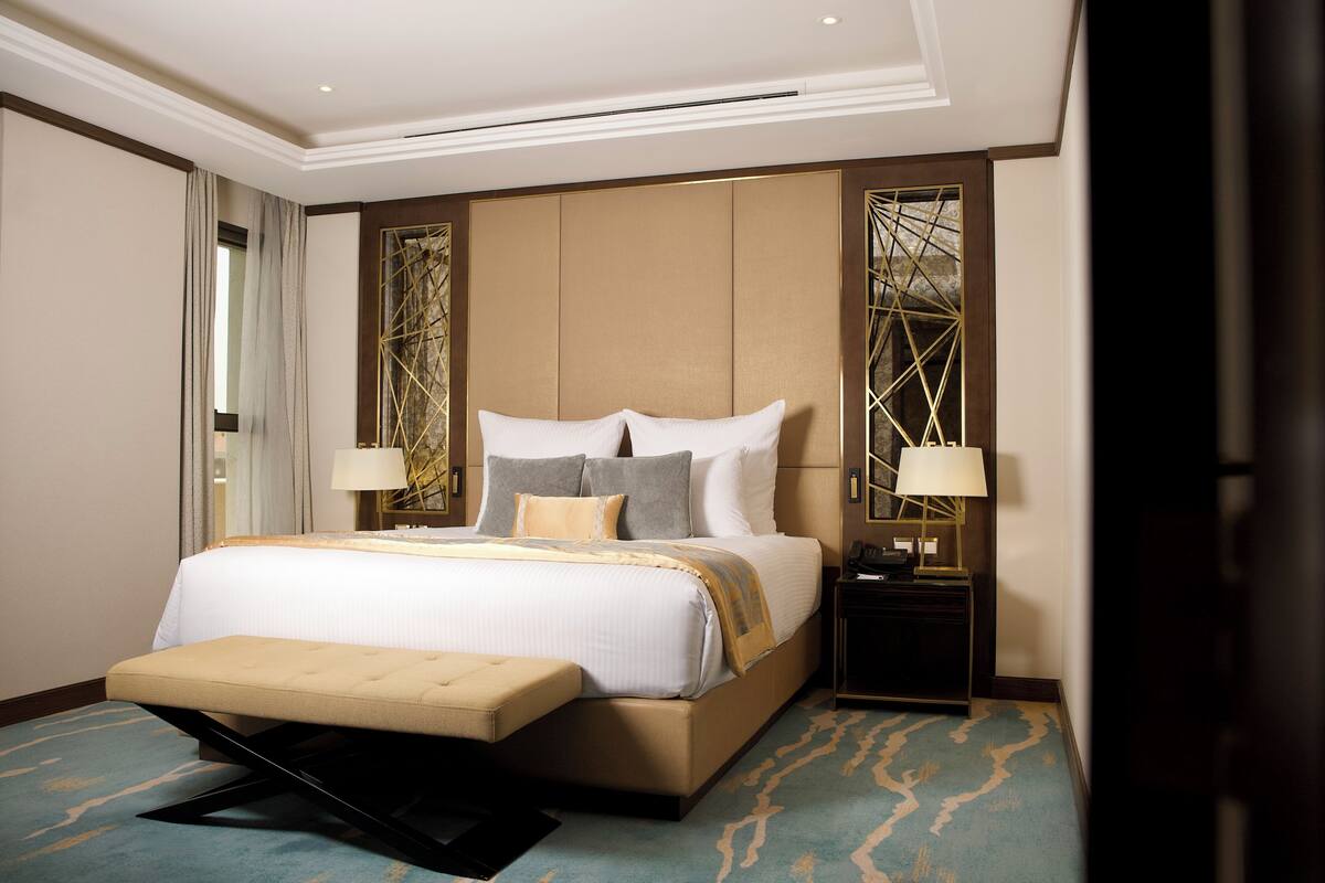 A spacious hotel room with a large bed, a bedside table with a lamp, and a decorative wall panel.