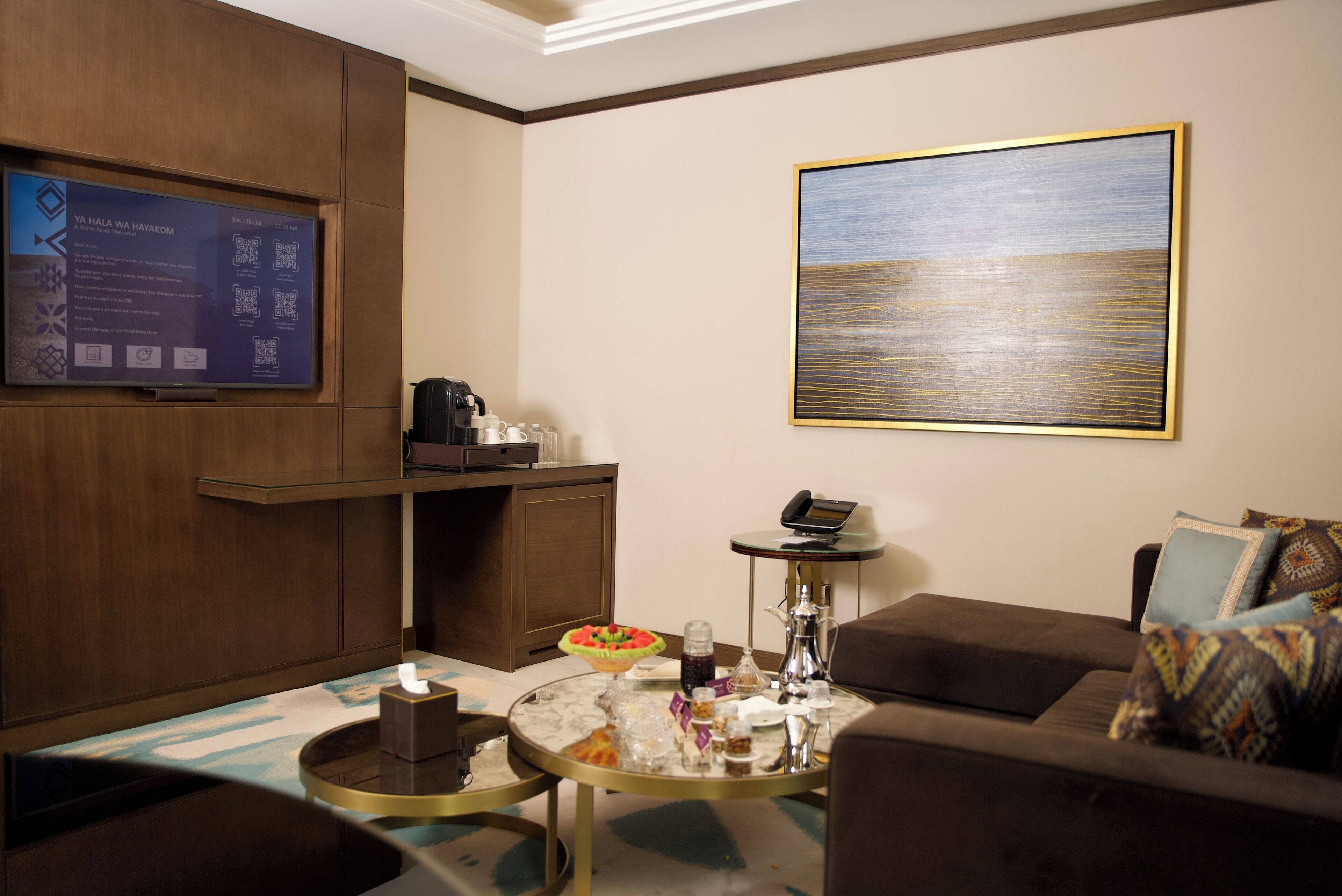 A modern hotel room with a large flat-screen TV mounted on the wall, a coffee machine, a small table with a bowl of fruit, and a sofa with patterned cushions.