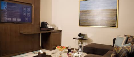 Une chambre d’hôtel moderne dotée d’un grand téléviseur à écran plat fixé au mur, d’une machine à café, d’une petite table avec un saladier de fruits et d’un canapé agrémenté de coussins à motifs.