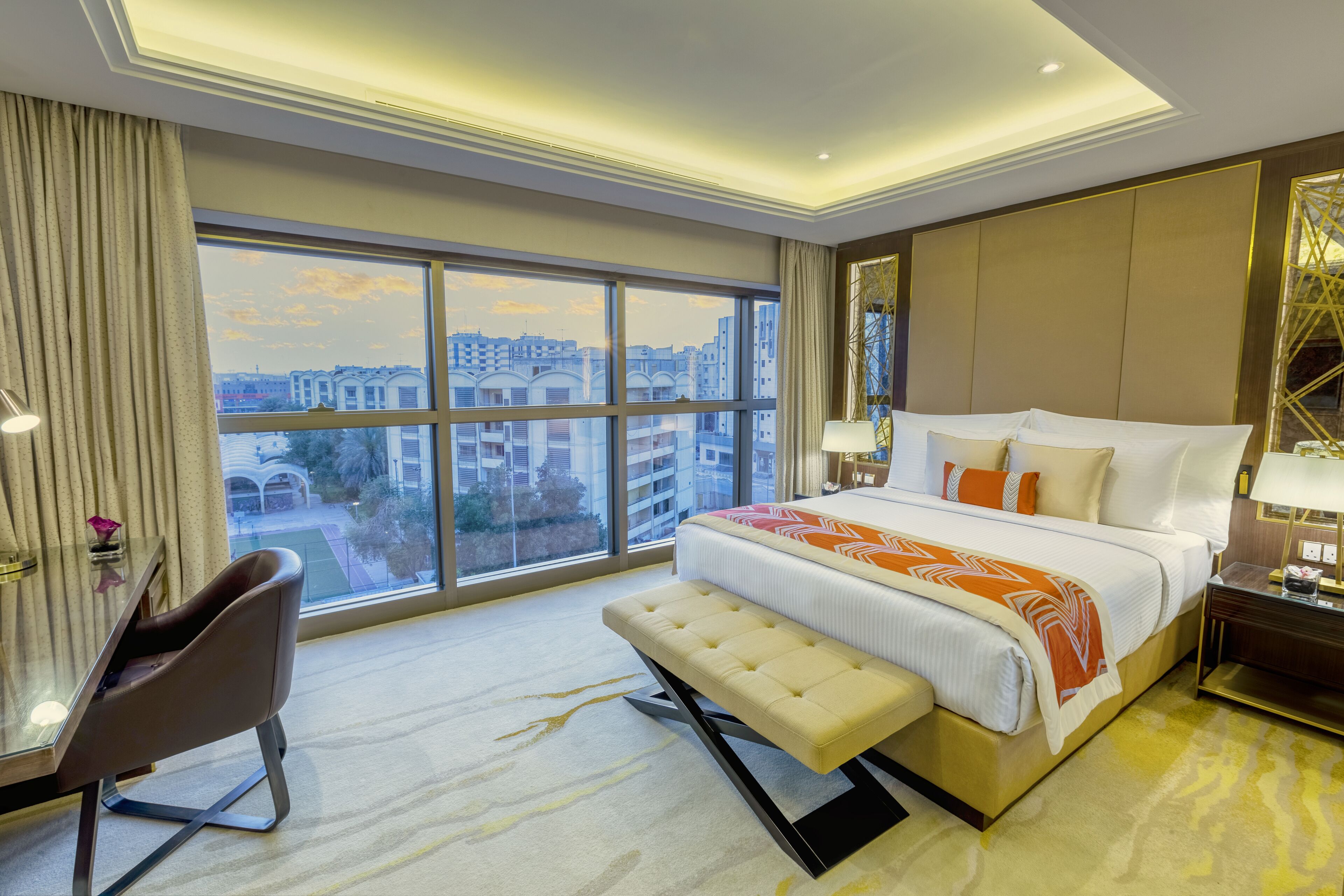 Habitación de hotel con una cama grande, una banca, un escritorio y vistas a la ciudad a través de amplias ventanas.