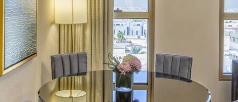 A modern hotel room with a round glass table, two grey upholstered chairs, a large framed artwork, a floor lamp, and a view of the city through large windows.