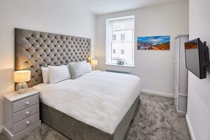 A bedroom with a tufted headboard, a large bed with white linens, a bedside table with a lamp, a window with blinds, a painting on the wall, and a TV on the wall. - Host & Stay | The Connell (Whitby)
