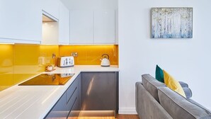 A modern kitchen with white cabinets, a yellow backsplash, and a gray sofa with a painting above it. - Host & Stay | The Connell (Whitby)