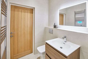 A modern bathroom with a wooden door, a white toilet, a white sink with a mirror above it, and a towel rack to the left. - Host & Stay | The Connell (Whitby)