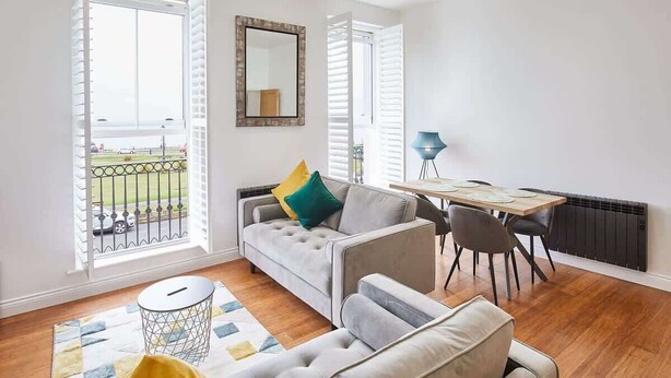 A modern living room with a grey sofa, a dining table with chairs, a small round table with a lamp, and a view of a balcony with a railing. - Host & Stay | The Connell (Whitby)