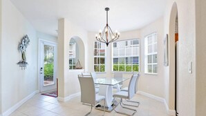A spacious dining area with a glass table and white chairs, a chandelier, and large windows with blinds. - Casa Boho | Cape Harbour 3/2 Coach Home (Cape Coral)