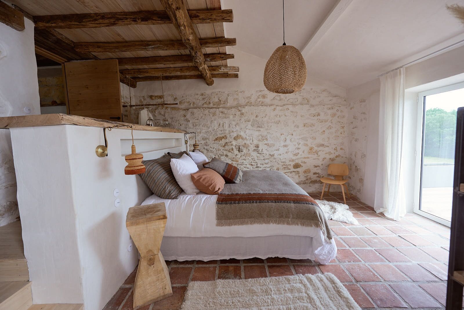 A rustic bedroom with exposed wooden beams, a stone wall, a bed with multiple pillows, a hanging light fixture, and a view through a large window.