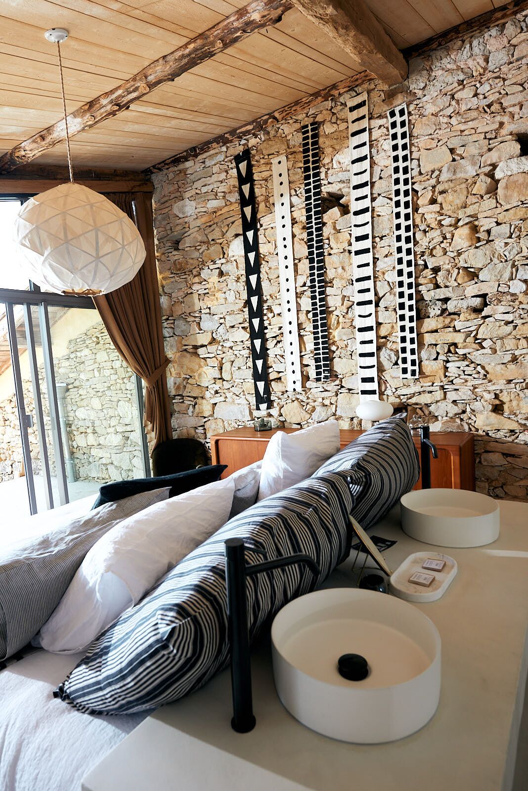 A bedroom with a stone wall, a bed with striped pillows, a hanging light fixture, and a bedside table with a lamp.