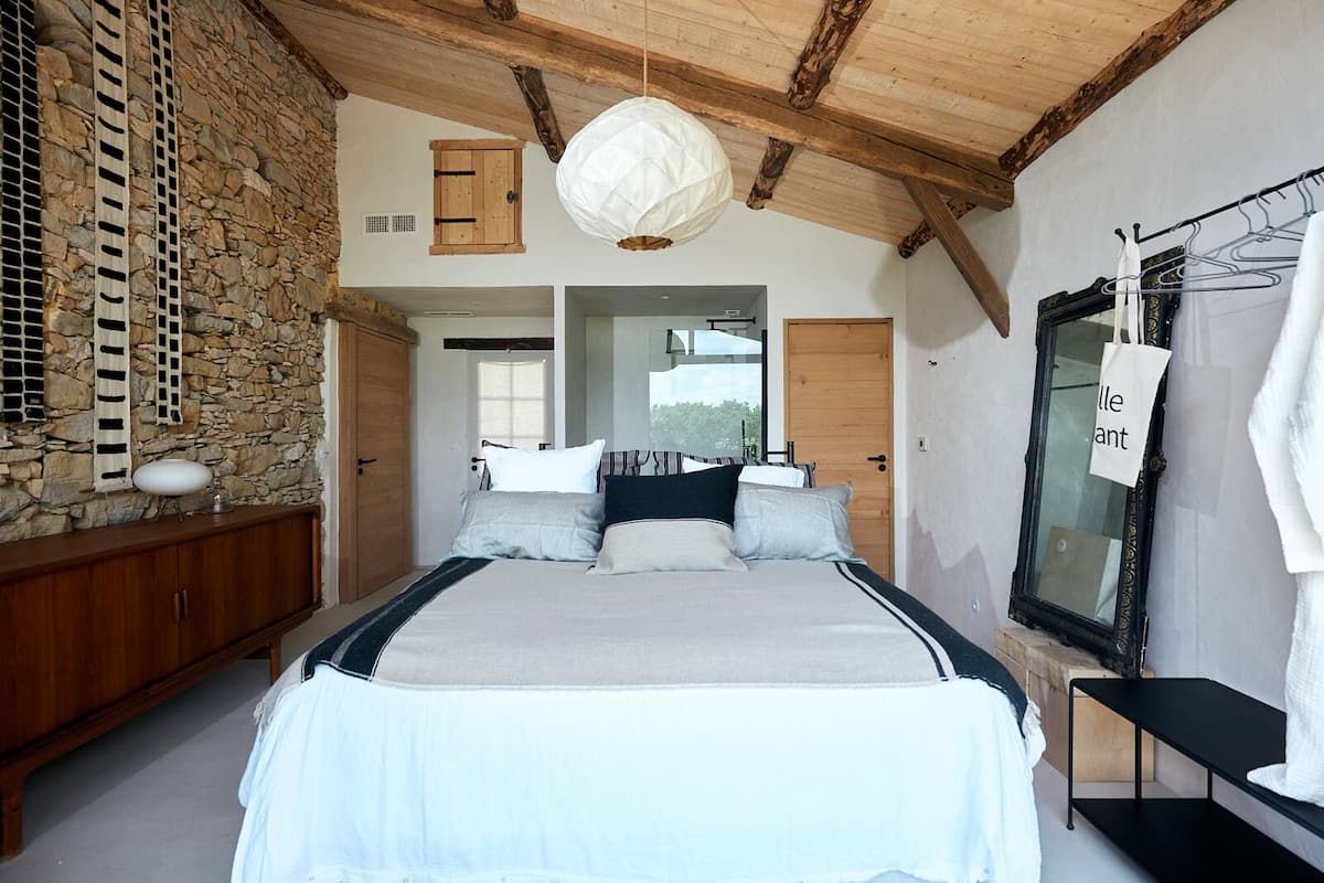 Un dormitorio con una cama grande, pared de piedra, vigas de madera y una lámpara colgante.