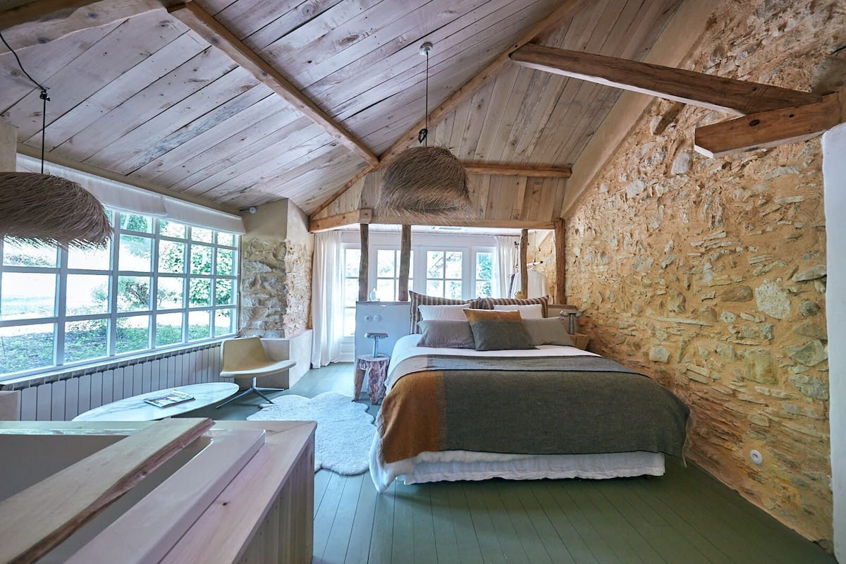 Un dormitorio rústico con pared de piedra, vigas de madera en el techo, una cama grande, un escritorio y una silla.