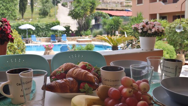 Un tavolo con croissant, fragole, uva e tazze di caffè, con vista su una piscina e vegetazione.
